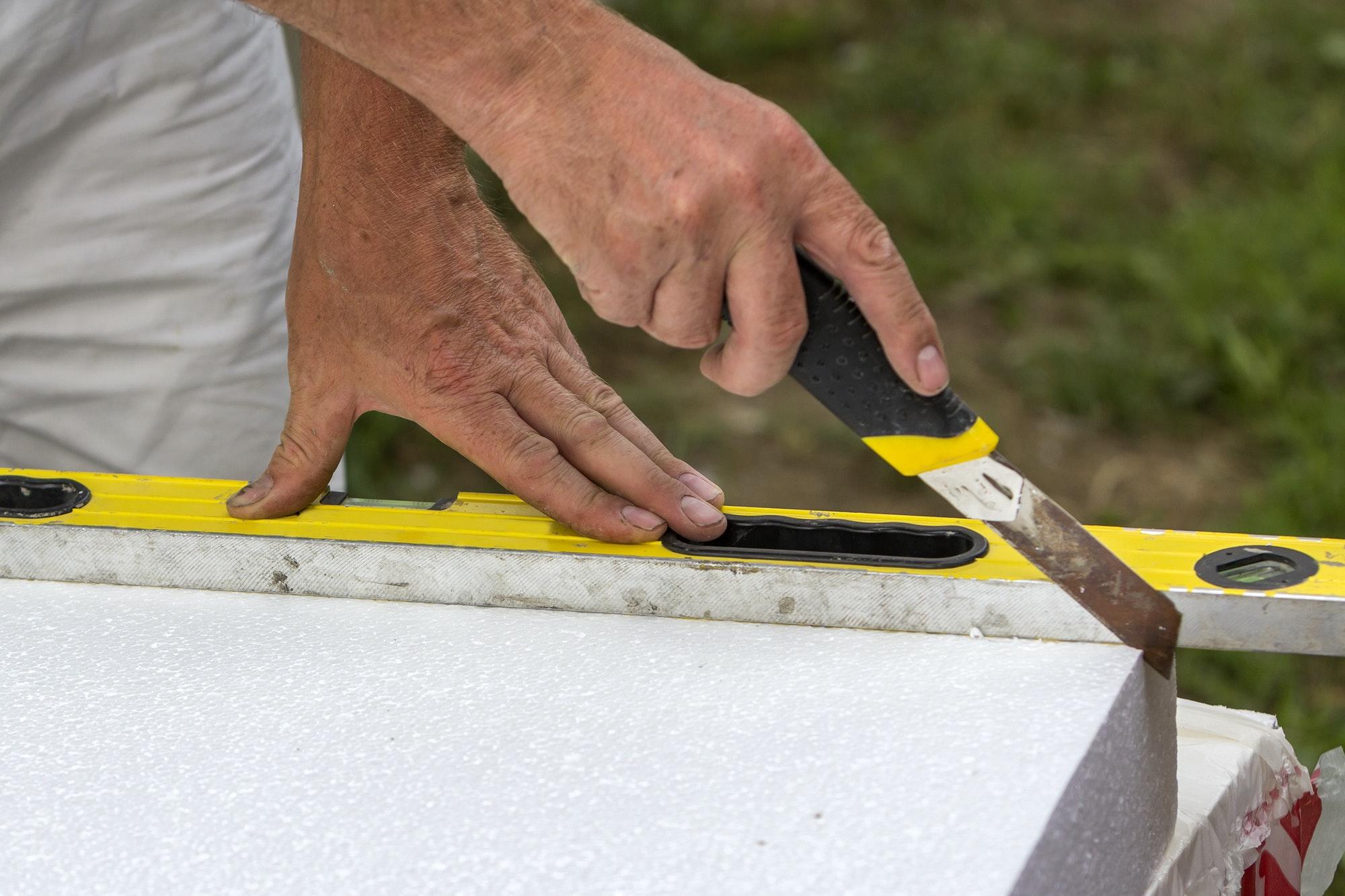 Close-up of worker hand with knife and level cutting white rigid polyurethane foam sheet for house